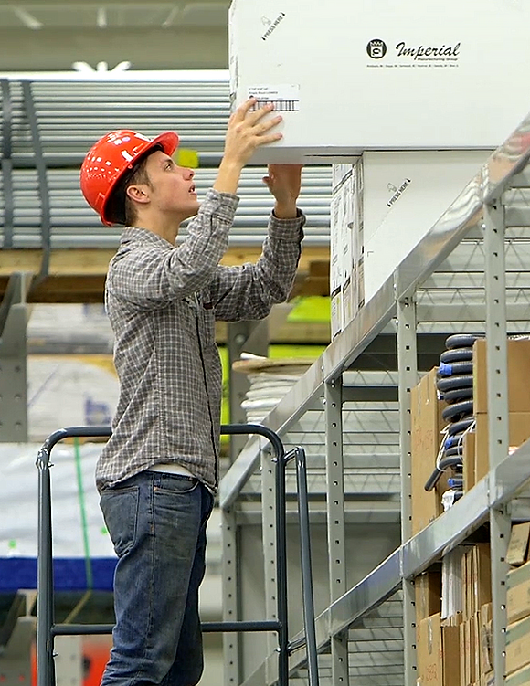 A person in a red hard hat and plaid shirt stands on a ladder, reaching for a box on a high shelf in a warehouse. The scene conveys focus and diligence.