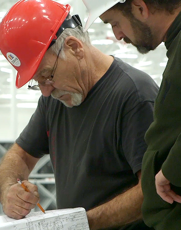 Two construction workers, one in a red hard hat, the other in white, review blueprints. Focused expressions suggest collaboration.