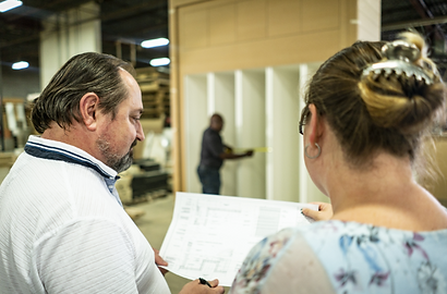 Two individuals review architectural plans in a warehouse. A worker in the background measures built-in shelving. The atmosphere is focused and industrious.