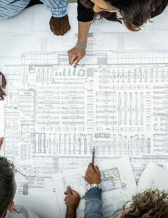 Four people collaborate over an architectural blueprint spread across a table. They are actively discussing and marking points with pens.