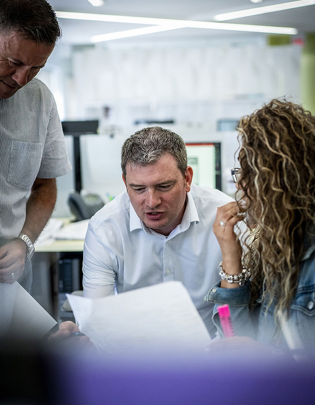 Three people in an office, two men and a woman, closely examining documents. They appear engaged and focused, conveying a collaborative atmosphere.