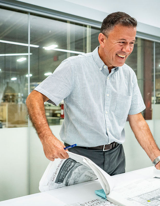 A smiling man in a light blue short-sleeve shirt leans over a table with blueprints. He stands in a bright office with glass walls, holding a pen.