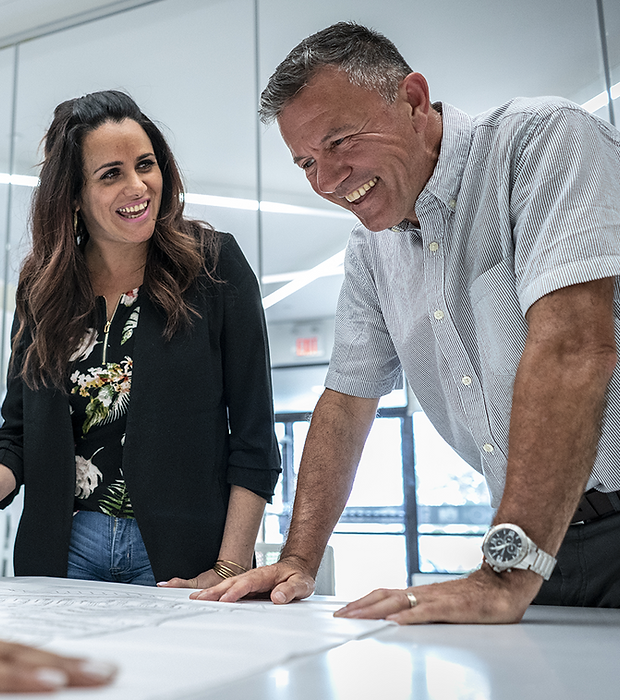 A man and woman are smiling and leaning over a table, reviewing documents in a bright office. They appear engaged and collaborative.