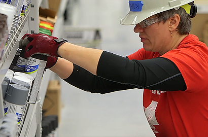A person wearing a hard hat and safety glasses stocks shelves with paint cans in a store. They wear a red shirt and gloves, focused and concentrated.